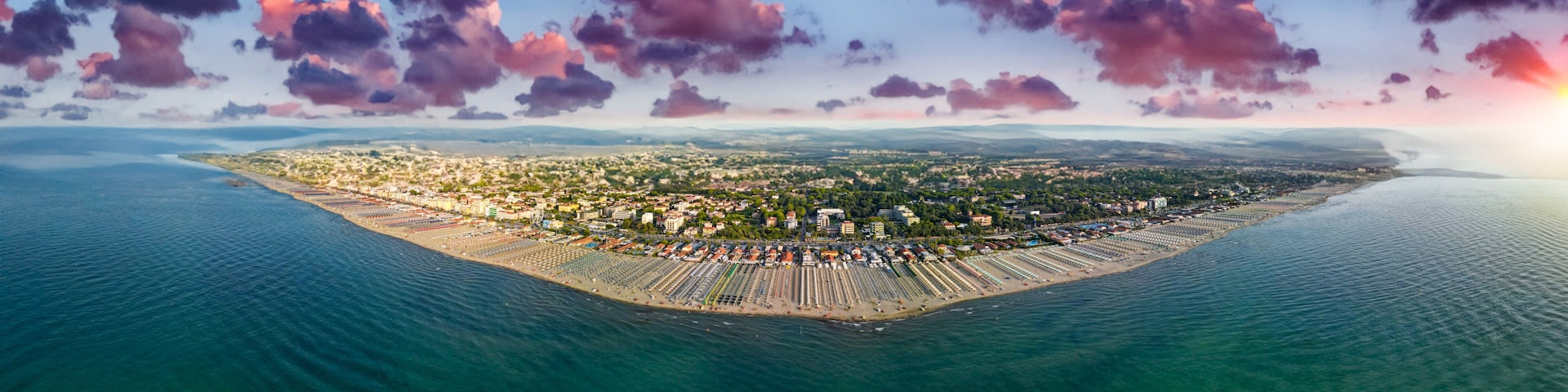 An aerial view of a coastal city with a densely populated urban area, a long beachfront lined with rows of beach umbrellas and sunbeds, and a backdrop of mountains and a sunset sky.