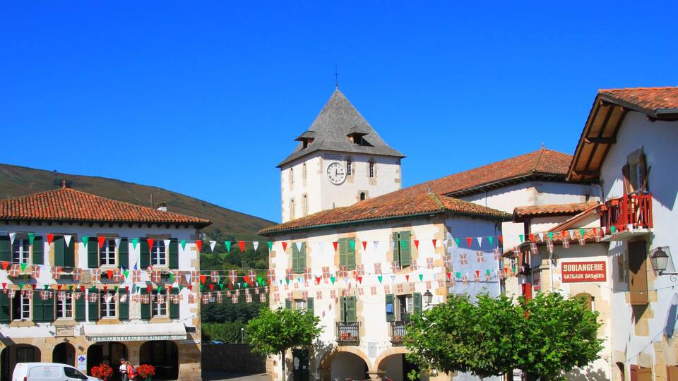 A town square with a clock tower, buildings with red-tiled roofs, and colorful flags.