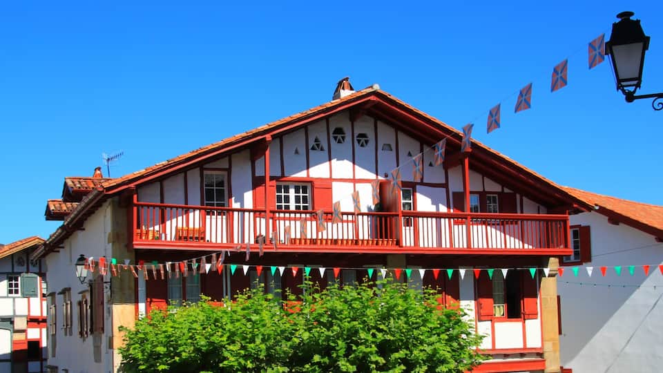 A traditional half-timbered house with a red balcony, surrounded by greenery and under a clear blue sky.