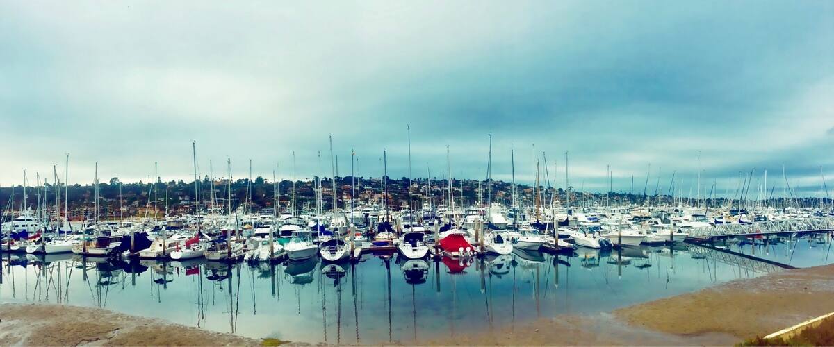 A marina with numerous boats docked, calm water, and a cloudy sky.