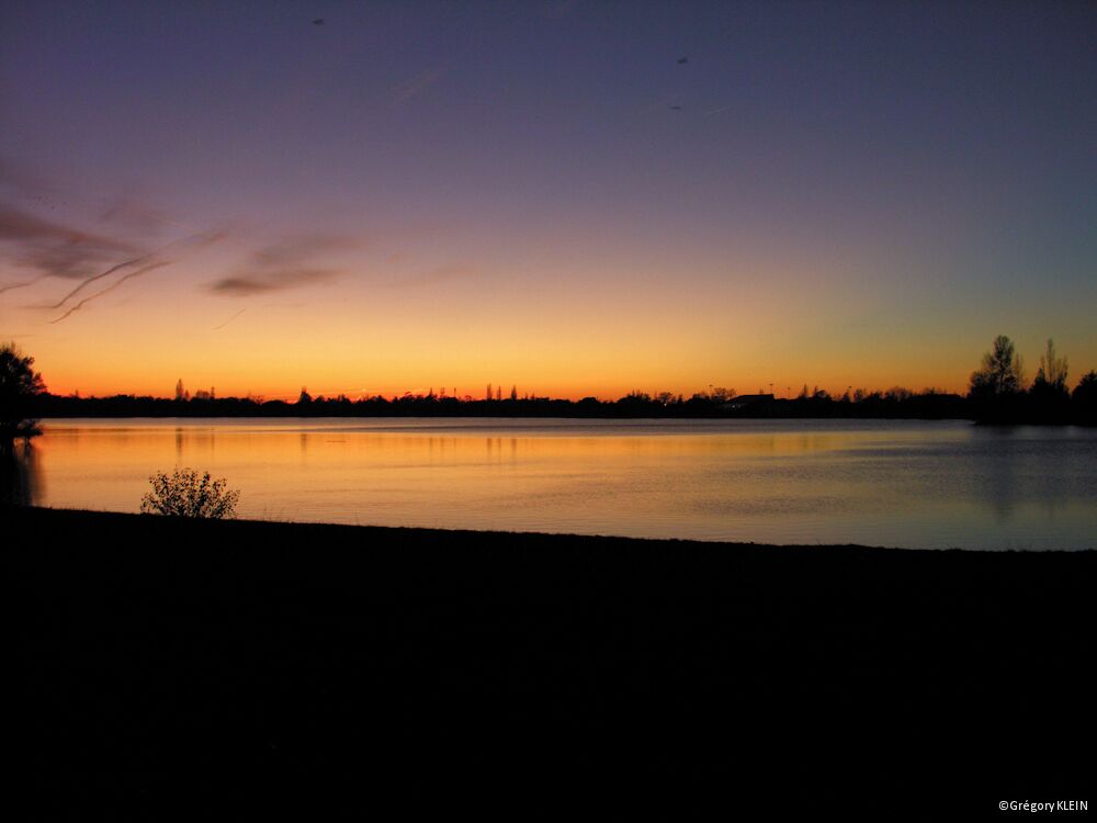 A sunset over a calm lake with a silhouette of trees and a distant city skyline.