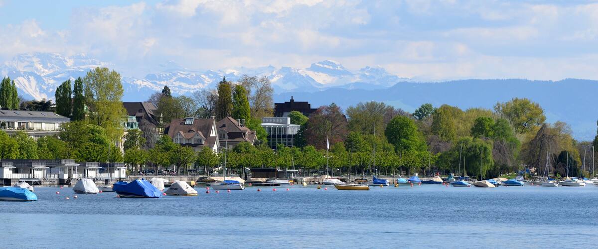 A lakeside town with boats, buildings, and snow-capped mountains in the background.