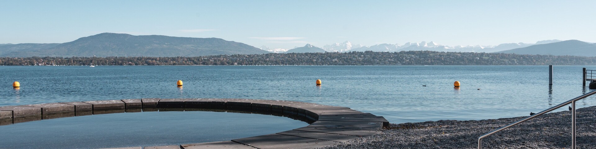 Uma praia de seixos com piscina e vista para montanhas ao longe.