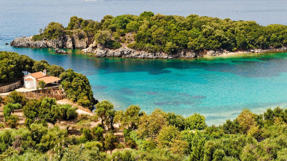 A coastal landscape with a small island covered in dense green trees, clear turquoise waters, and a white building on the shore.