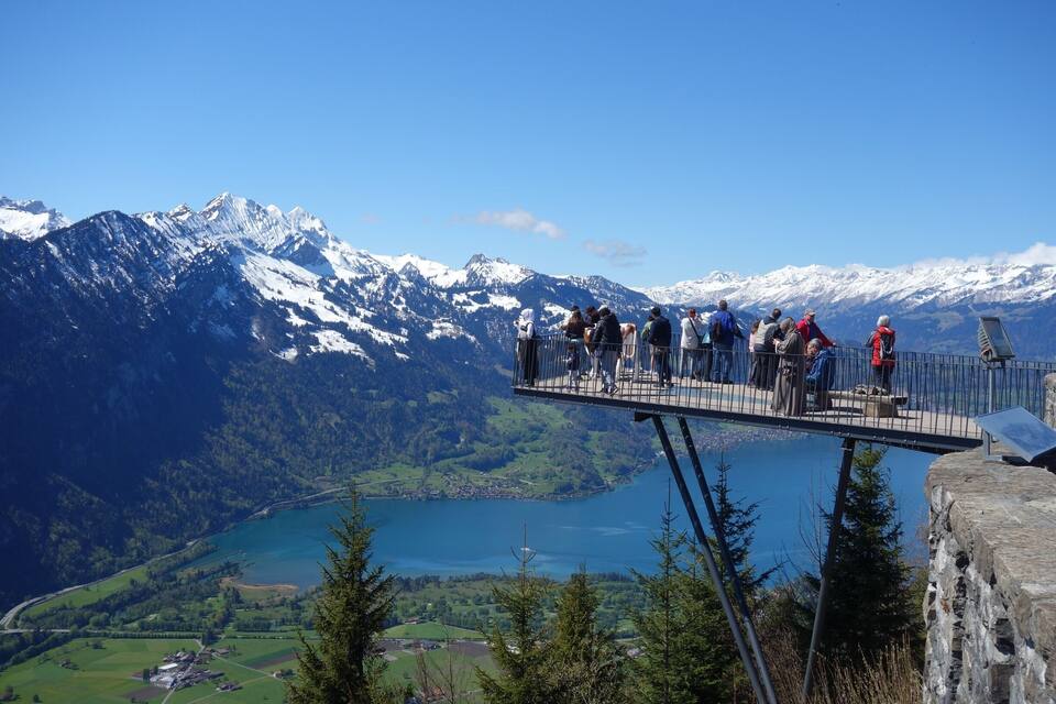Un grup de persones sobre un pont de vidre amb vista a un llac i muntanyes nevades.