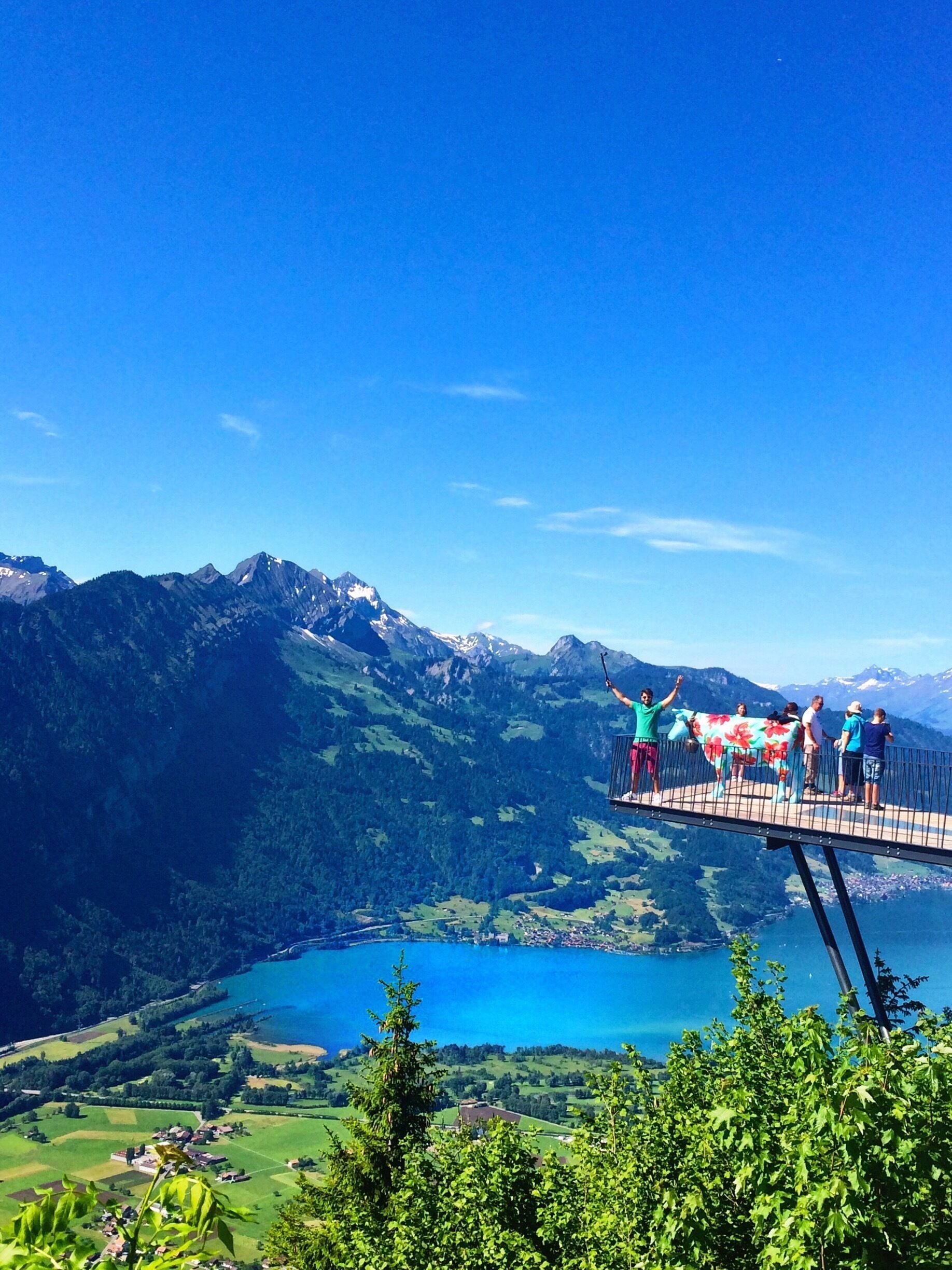 A scenic overlook with a lake, mountains, and a bridge with people enjoying the view.