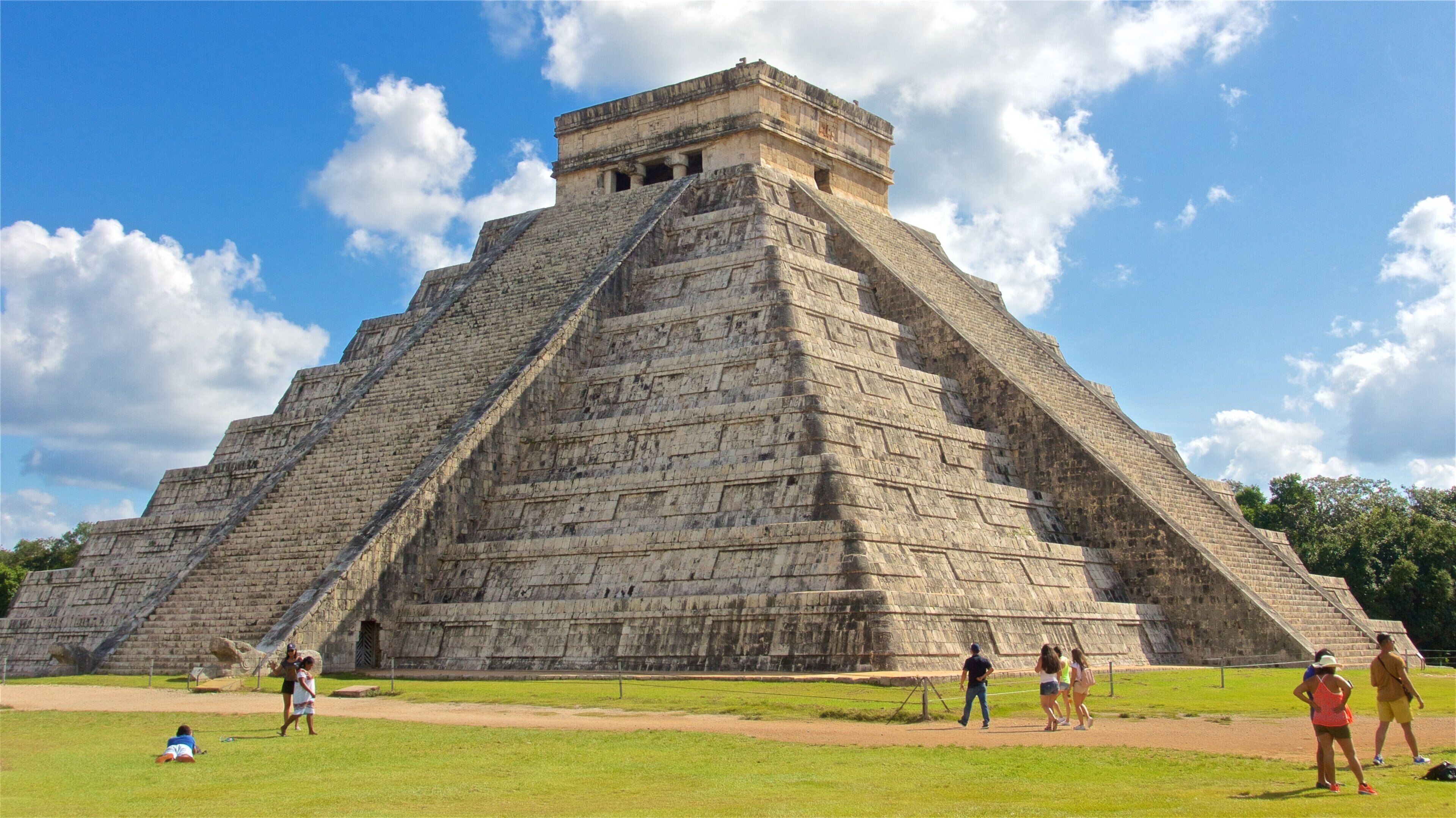 A large stone pyramid with a flat top, surrounded by a grassy area with people walking around.