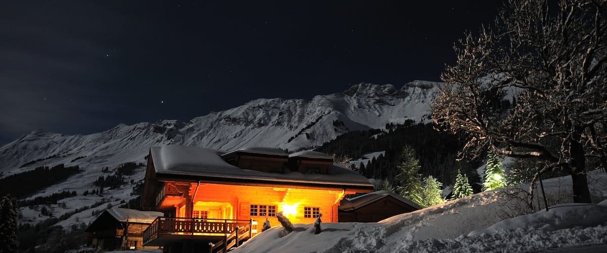 A snow-covered mountain with a lit chalet and a tree in the foreground.