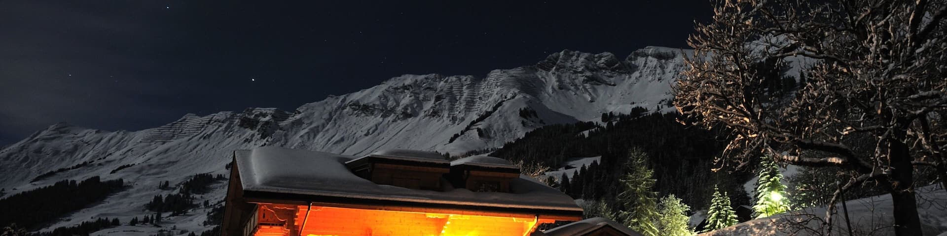 A snow-covered mountain with a lit chalet and a tree in the foreground.
