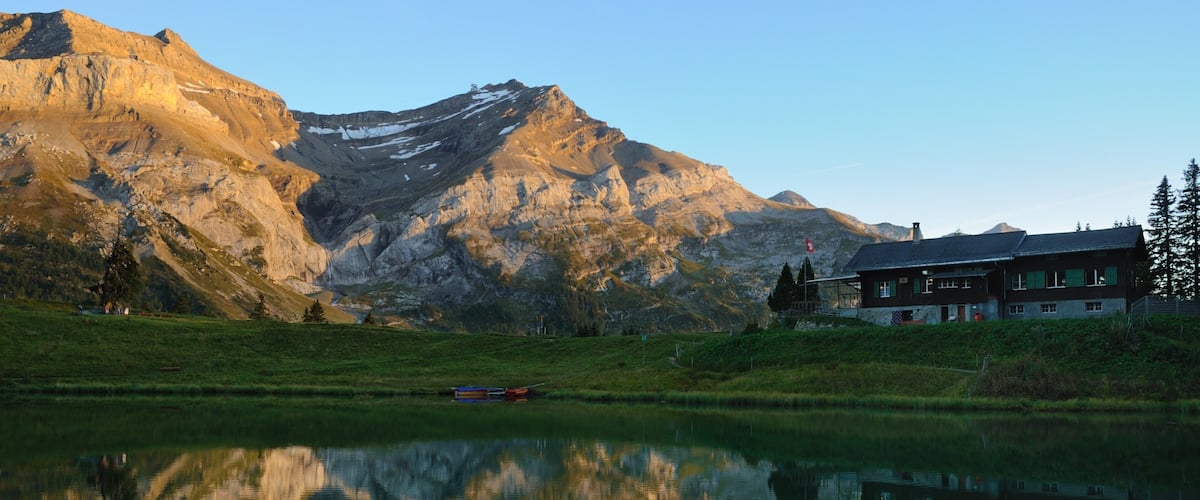 A mountain range with a lake reflecting the scenery, a house with a green roof, and a small boat on the water.