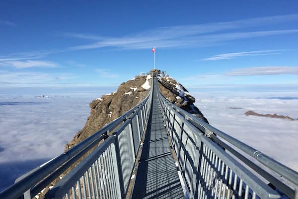 Un pont métallique menant au sommet d’une montagne, où flotte un drapeau, le tout entouré de nuages.