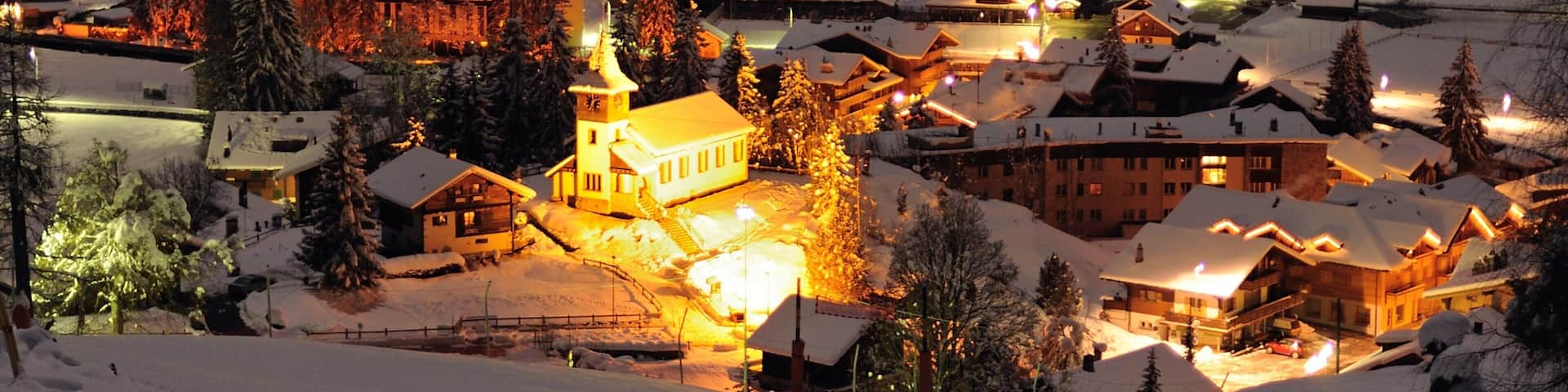 A snow-covered village at night with illuminated buildings and a church.