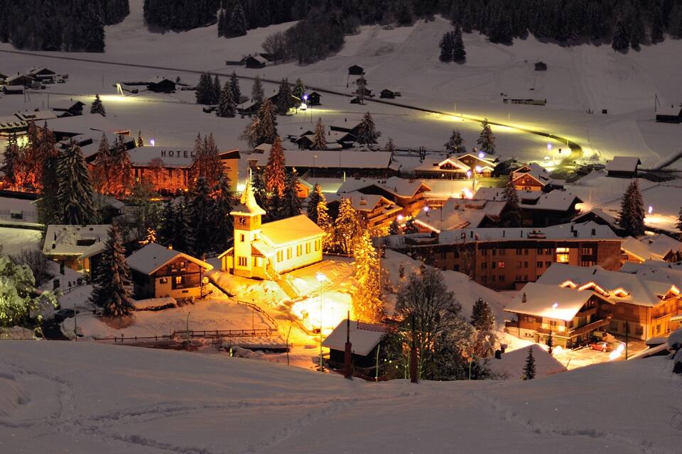 Un pueblo cubierto de nieve por la noche con edificios iluminados y una iglesia.