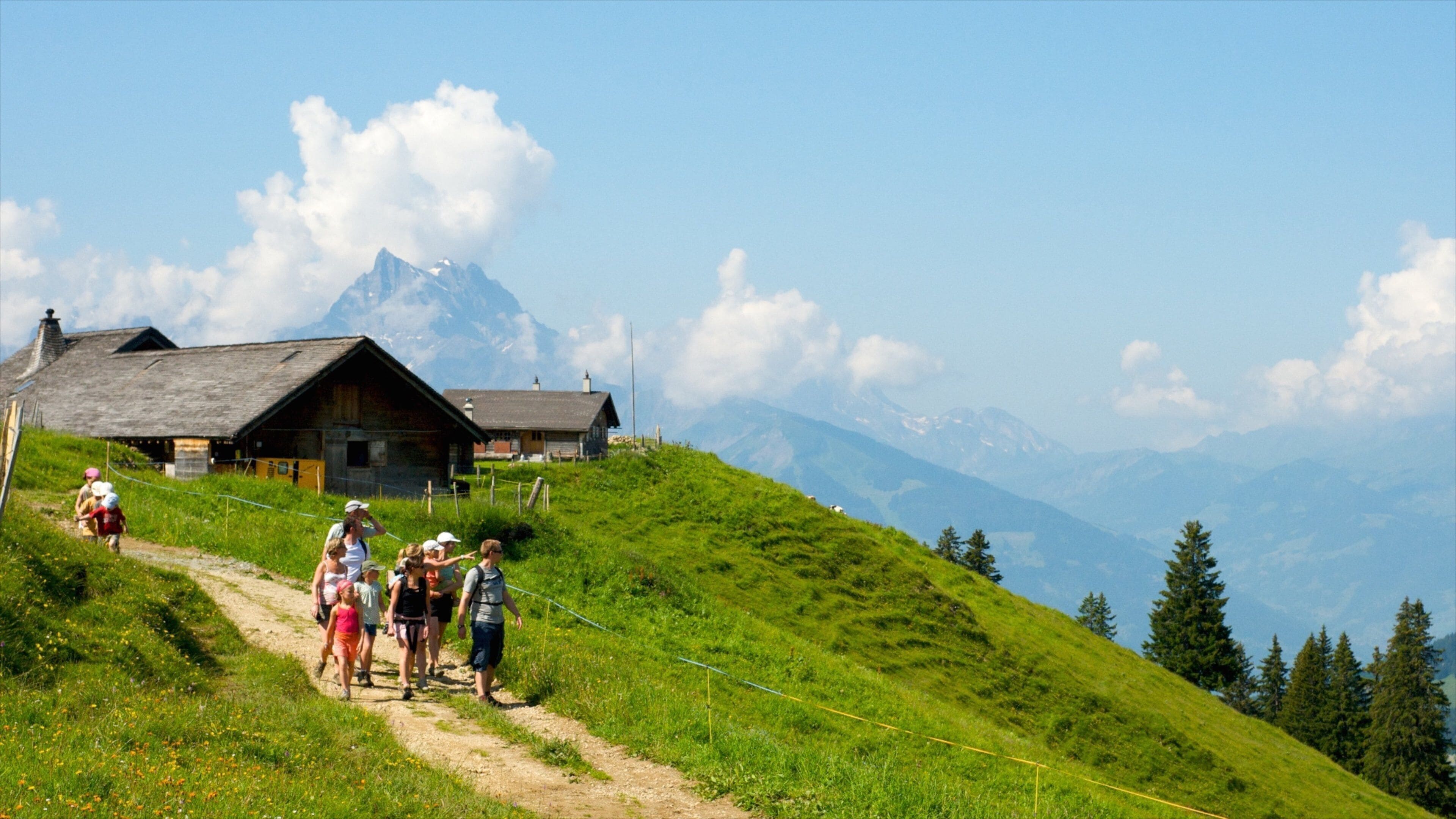 A group of hikers on a dirt path with a mountain in the background and a wooden house.