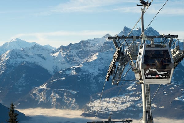 A chairlift with 'Villars-Gryon' branding, snow-covered mountains, and a forested valley in the background.