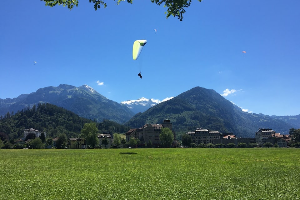 A grassy field with a person kite surfing, mountains in the background, and buildings near the field.