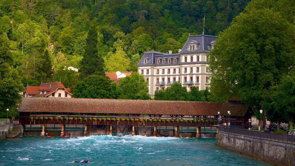 A riverside scene with a thatched-roof building, kayakers, and a large historic building in the background.