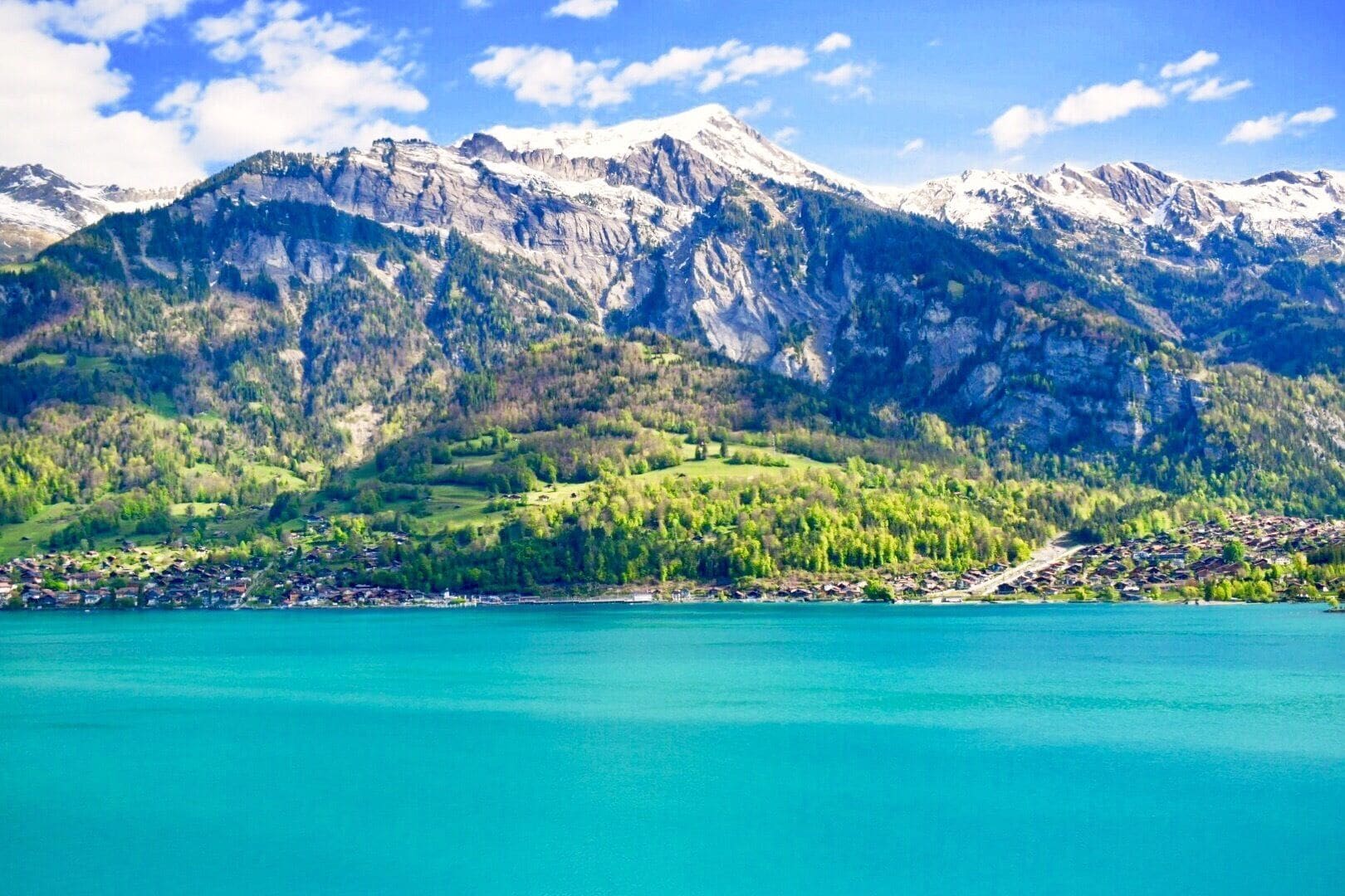 A mountain range with snow-capped peaks, a turquoise lake, and a village on the shore.