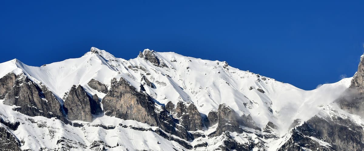 A snow-covered mountain range with sharp peaks and rocky outcrops under a clear blue sky.