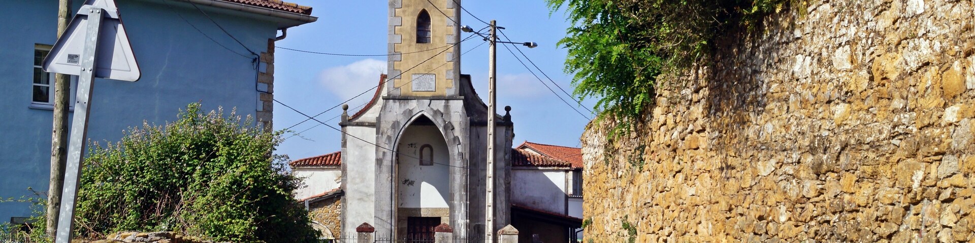 Uma estrada estreita que leva a uma igreja com um campanário, cercada por muros de pedra e vegetação.