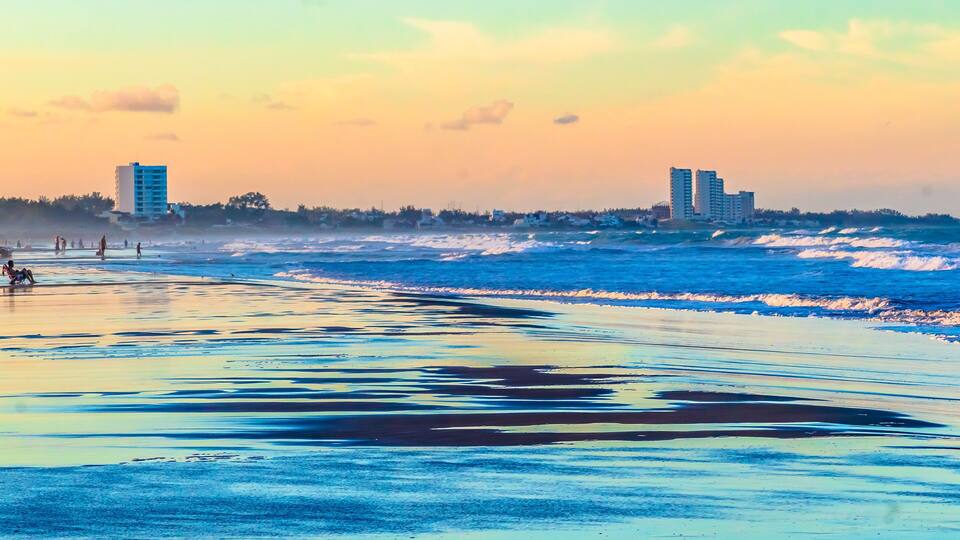 A beach with waves, a clear sky, and buildings in the background.