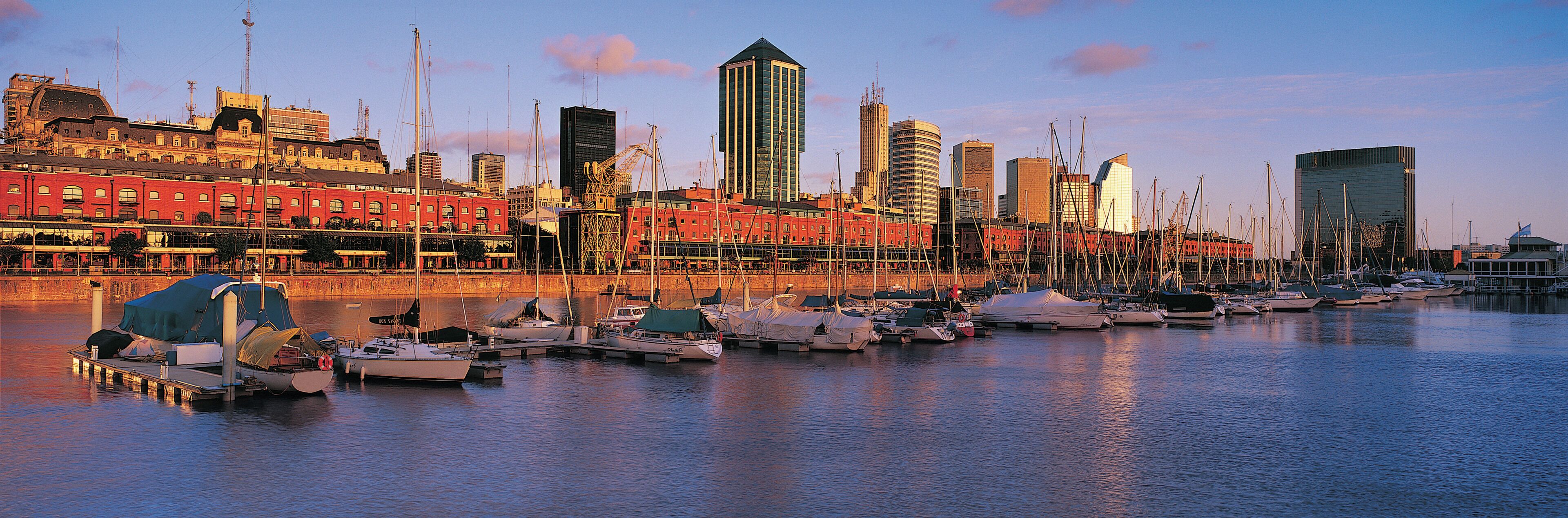 A marina with boats docked, a city skyline in the background, and a mix of modern and traditional architecture.