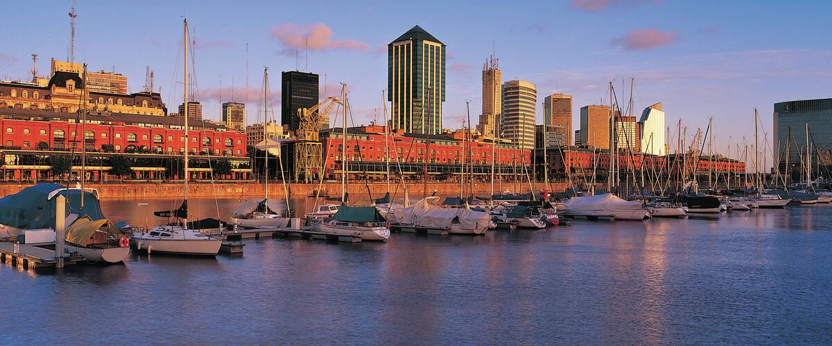A marina with boats docked, a city skyline in the background, and a mix of modern and traditional architecture.