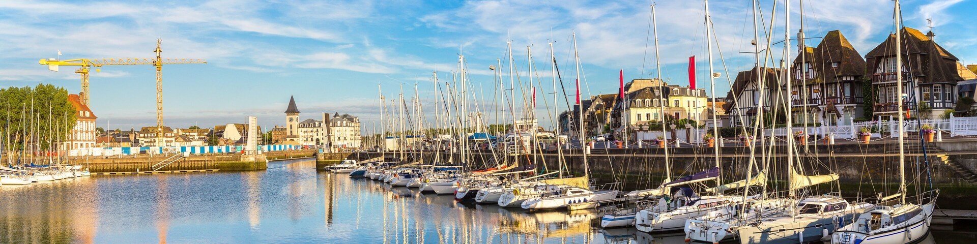 Un puerto deportivo con numerosos veleros amarrados, una grúa y edificios históricos al fondo.