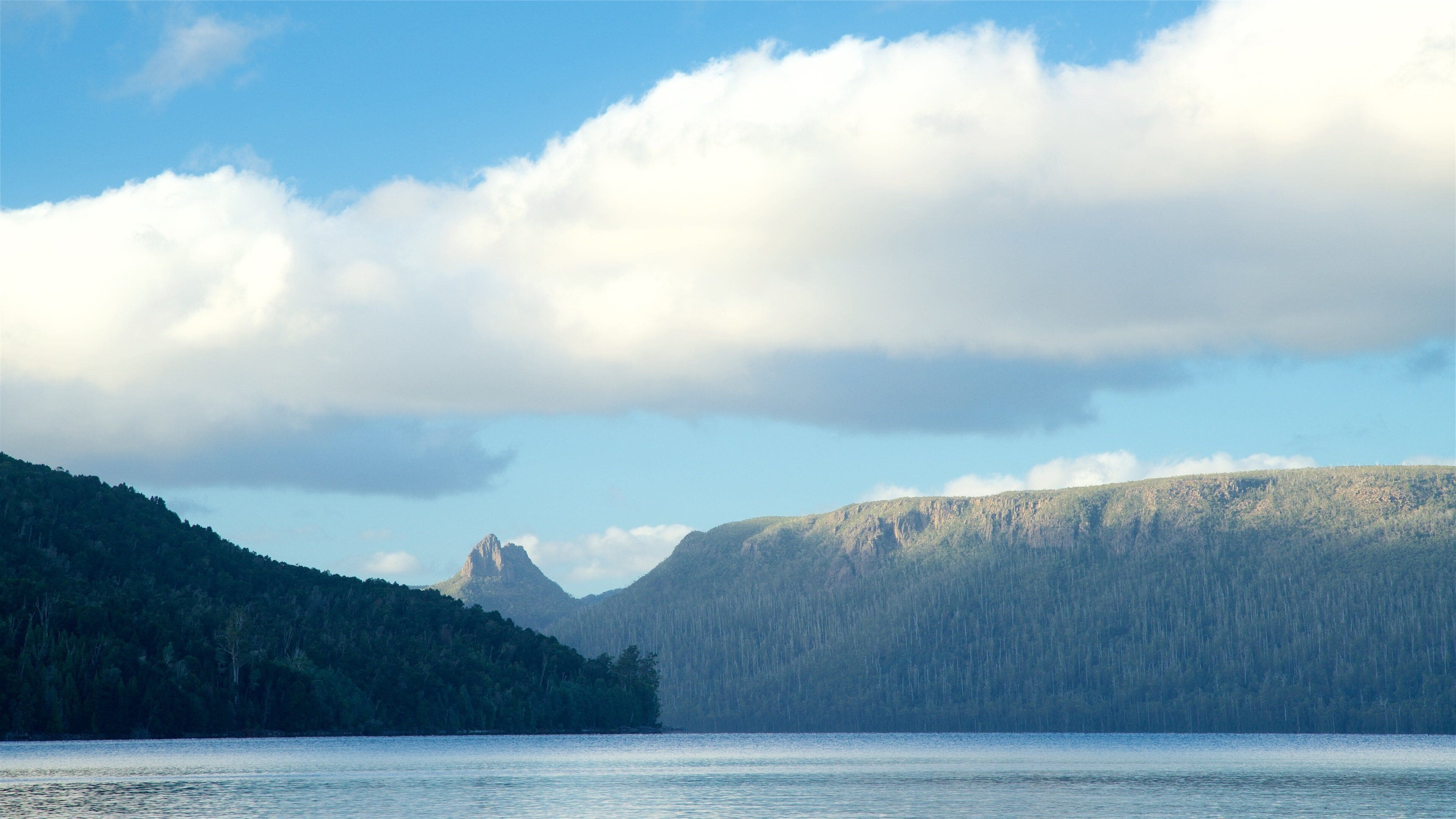 A calm lake with a forested mountain in the background and a prominent rocky peak.
