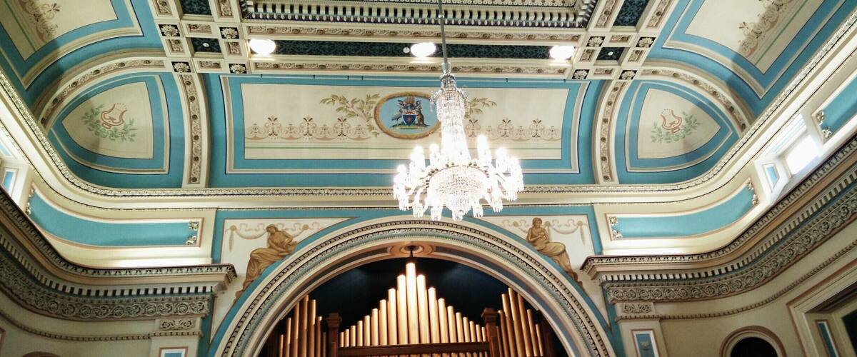 A grand hall with ornate blue and white ceilings, a large organ, and a chandelier.