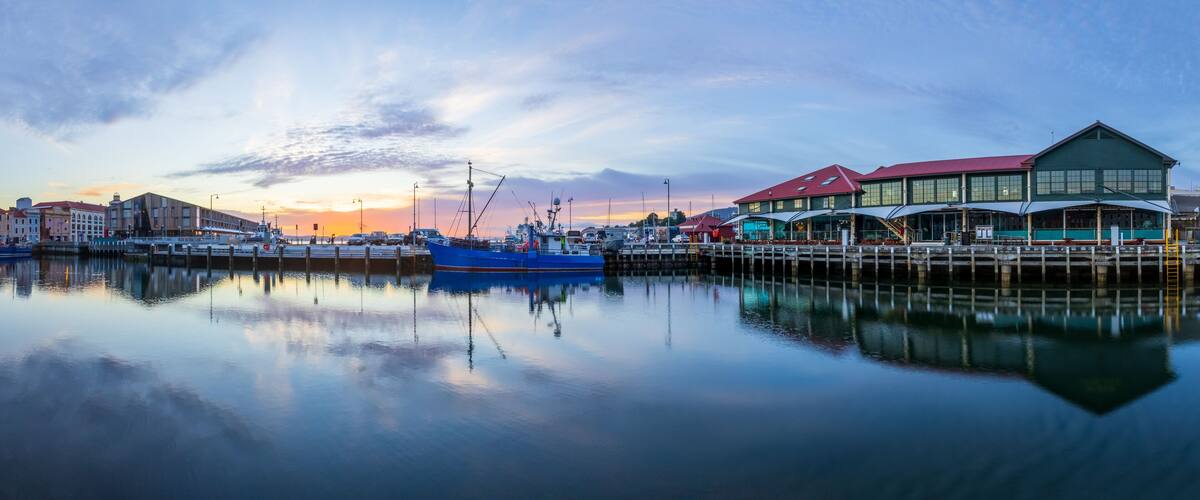 A harbor at sunset with boats, a pier, and buildings reflecting on the water.