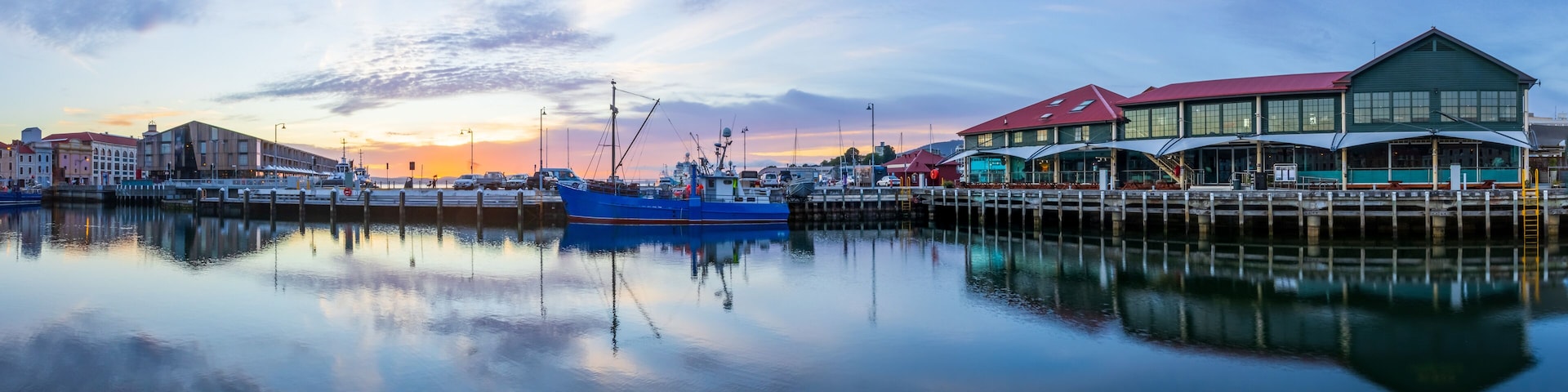 Un port au coucher du soleil, avec des bateaux, une jetée et des bâtiments dont le reflet se mire dans l’eau.