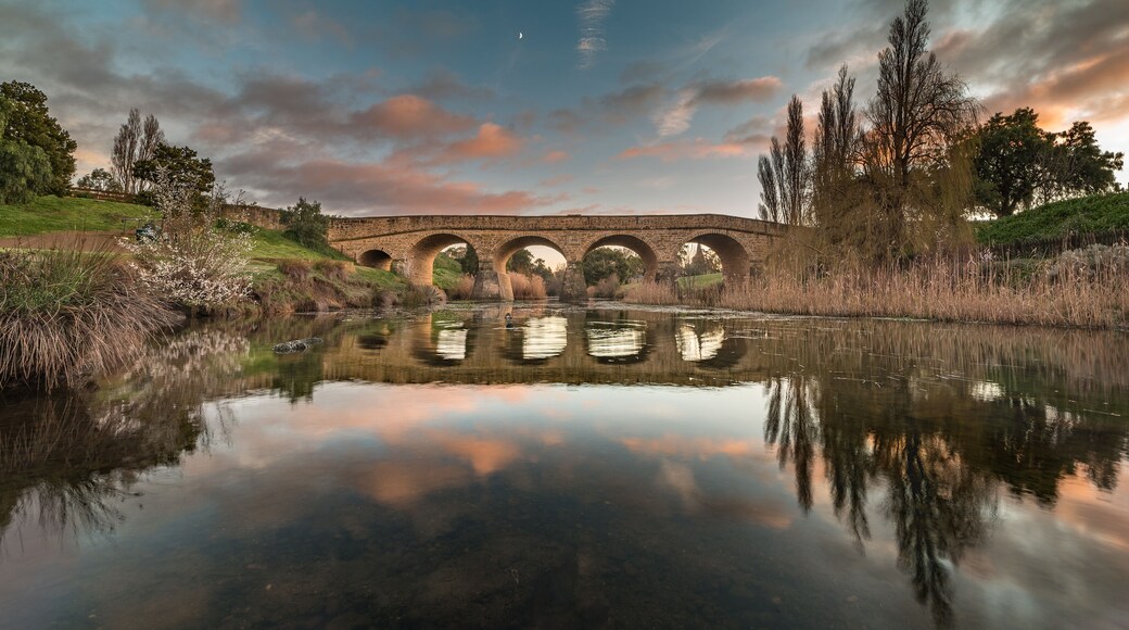 A stone bridge over a calm river with reflections, surrounded by trees and a clear sky.