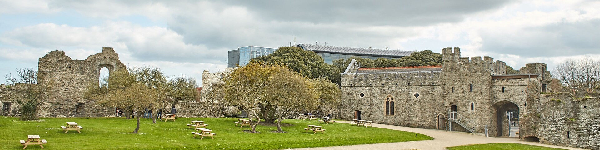 Um castelo histórico com ruínas, mesas de piquenique e um edifício moderno ao fundo.