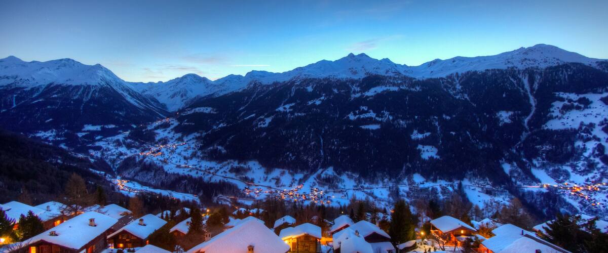 A snow-covered mountain range at dusk with illuminated houses in the foreground.