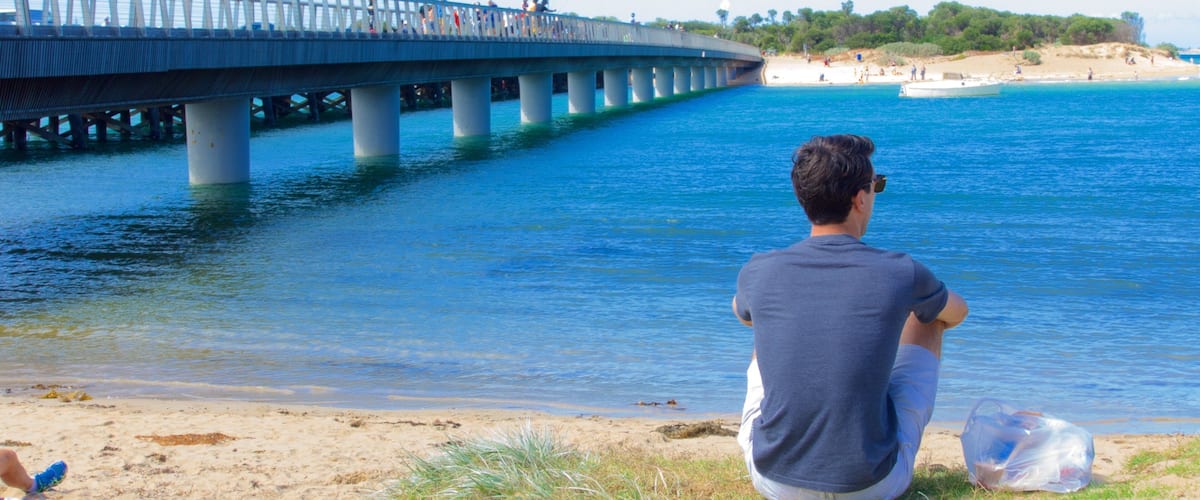 A man sitting on a beach with a pier extending into the water.