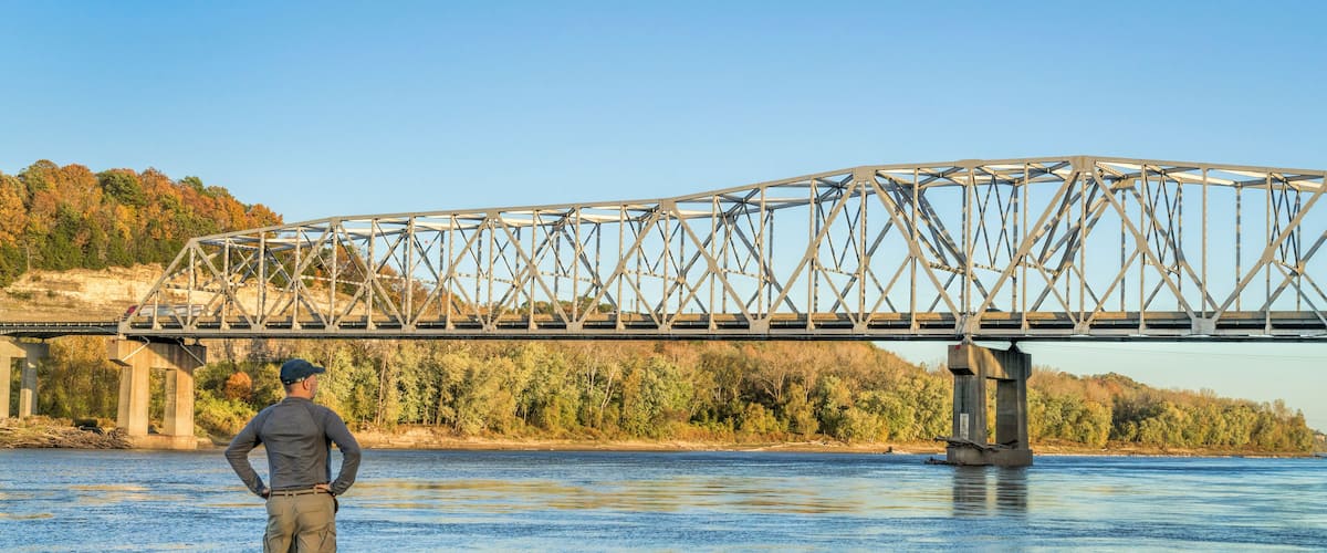 A man standing by a river with a large bridge in the background.