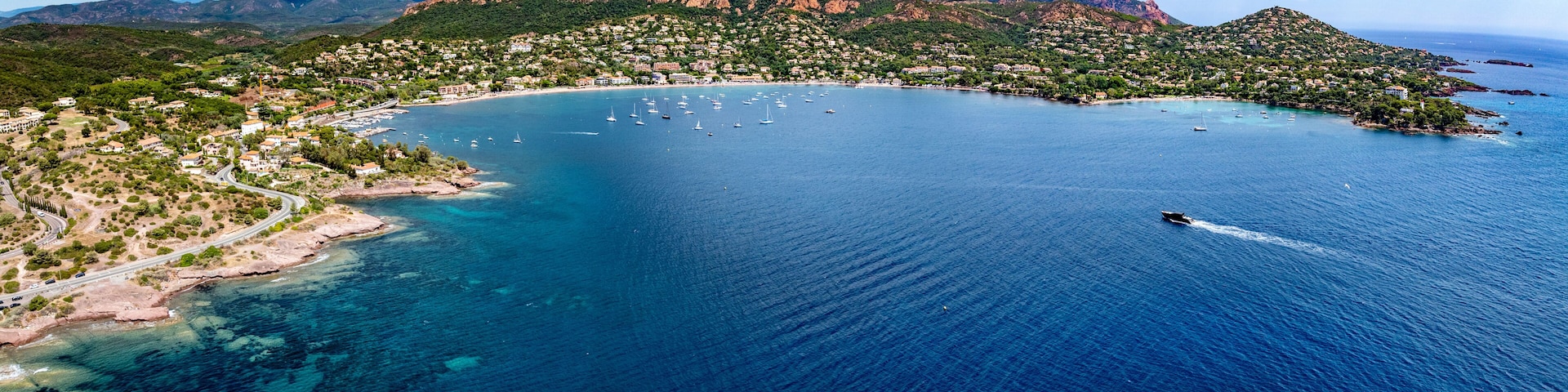 wide angle aerial landscape view of French Riviera coastline along Agay Bay in Saint-Raphaël with Plage d'Agay and Plage de La Baumette, boats anchored in bay, wooded hilly landscape in background