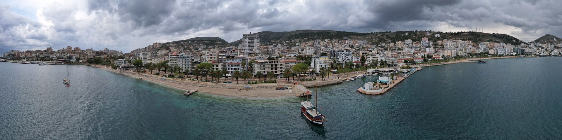 Un paisaje urbano costero con un puerto deportivo, edificios y un paisaje montañoso al fondo.