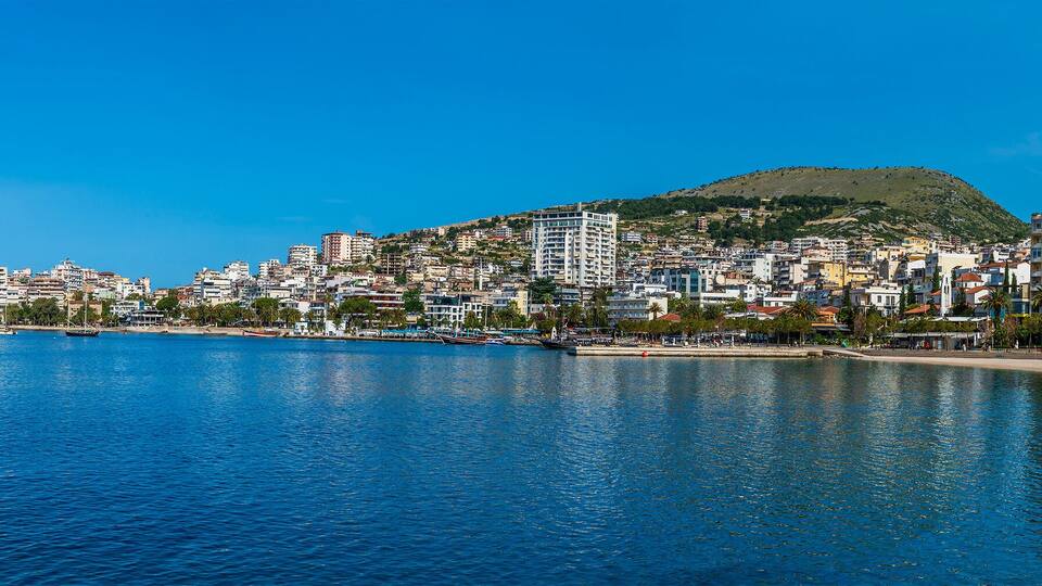 A coastal cityscape with a clear blue sea, a sandy beach, and a hilly background.