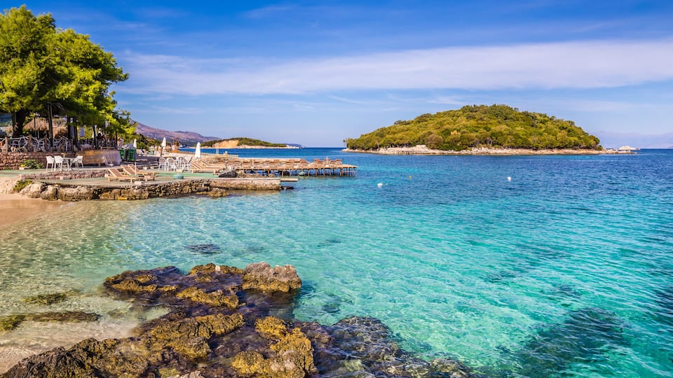 A clear turquoise sea, rocky shoreline, a pier with white chairs, and a small green island.