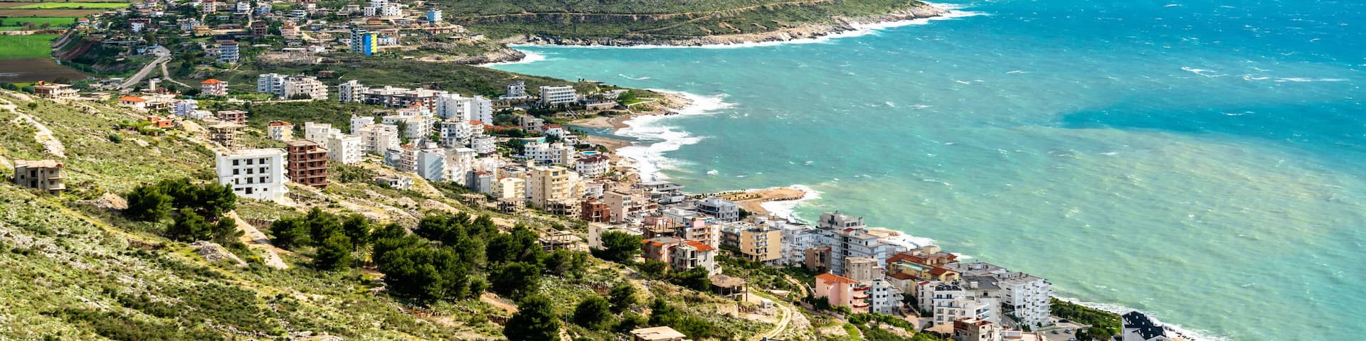 A coastal town with buildings along the shoreline, a clear view of the sea, and rolling hills in the background.