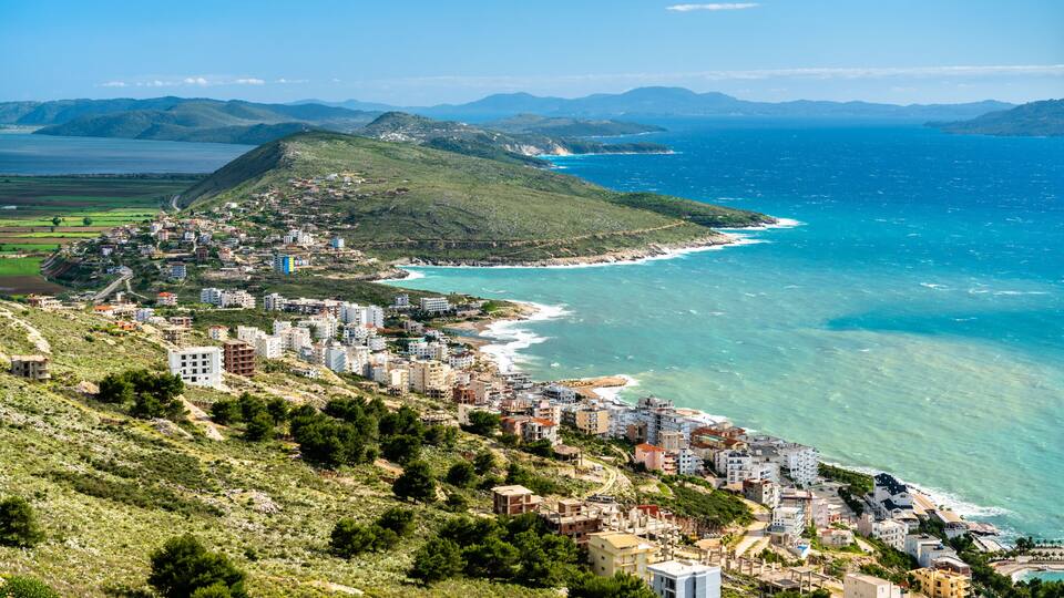 A coastal town with buildings along the shoreline, a clear view of the sea, and rolling hills in the background.