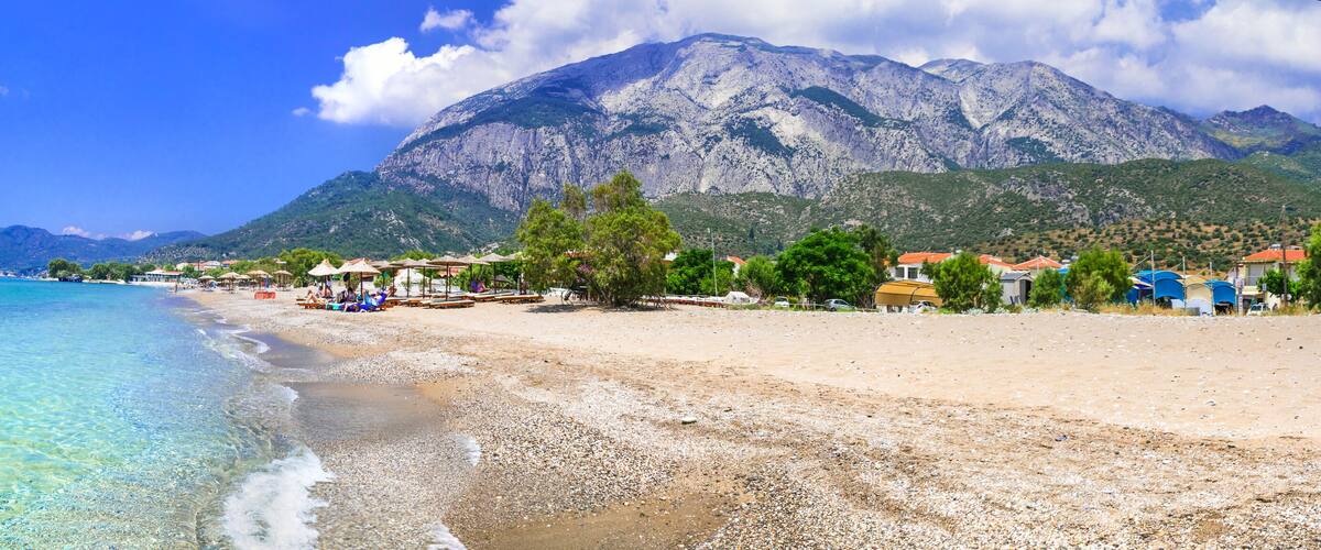 A beach with clear turquoise water, pebbly shore, and a mountain in the background.