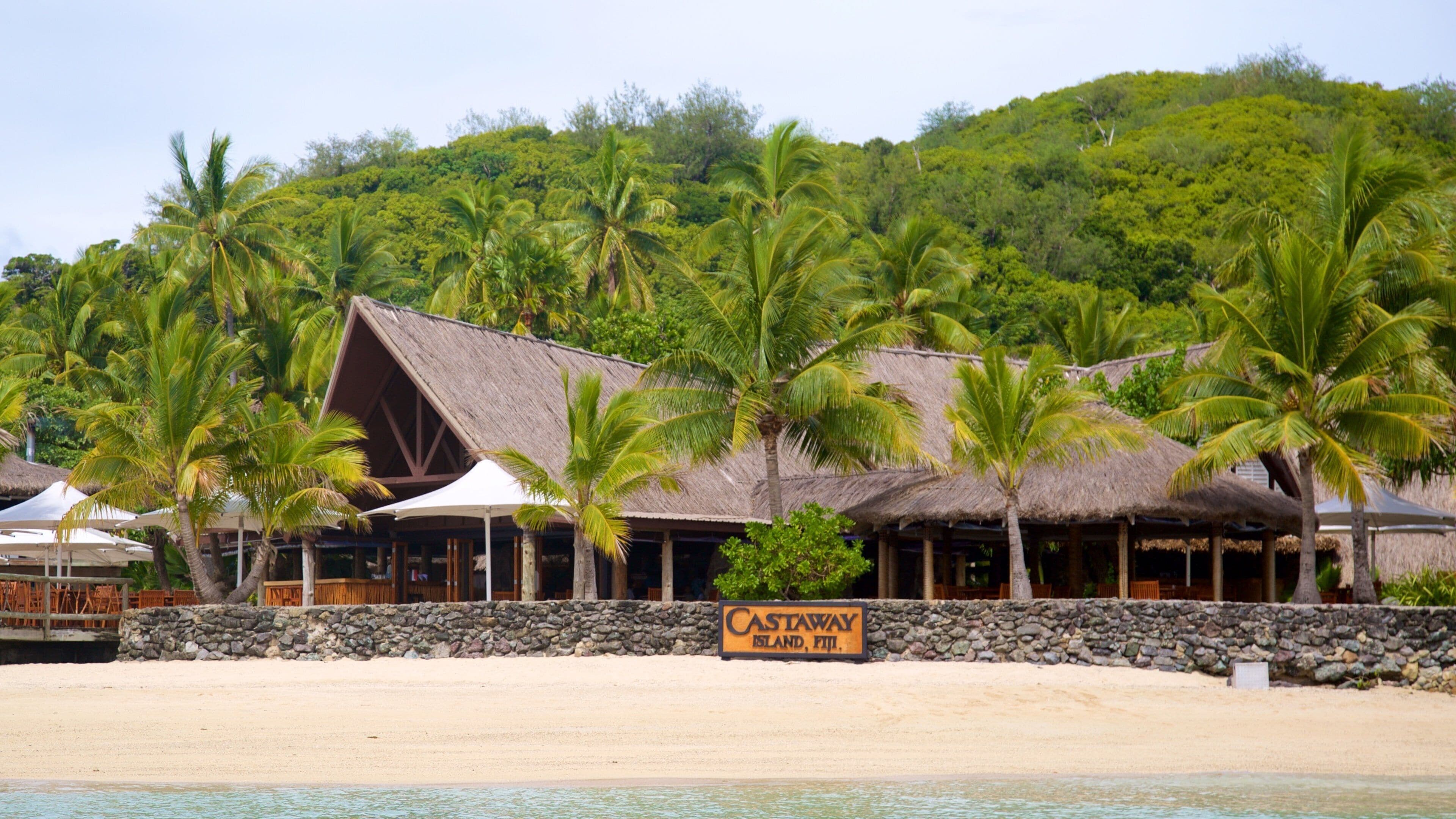 A beachside resort with thatched-roof buildings, palm trees, and a sign reading "Castaway Island, Fiji."