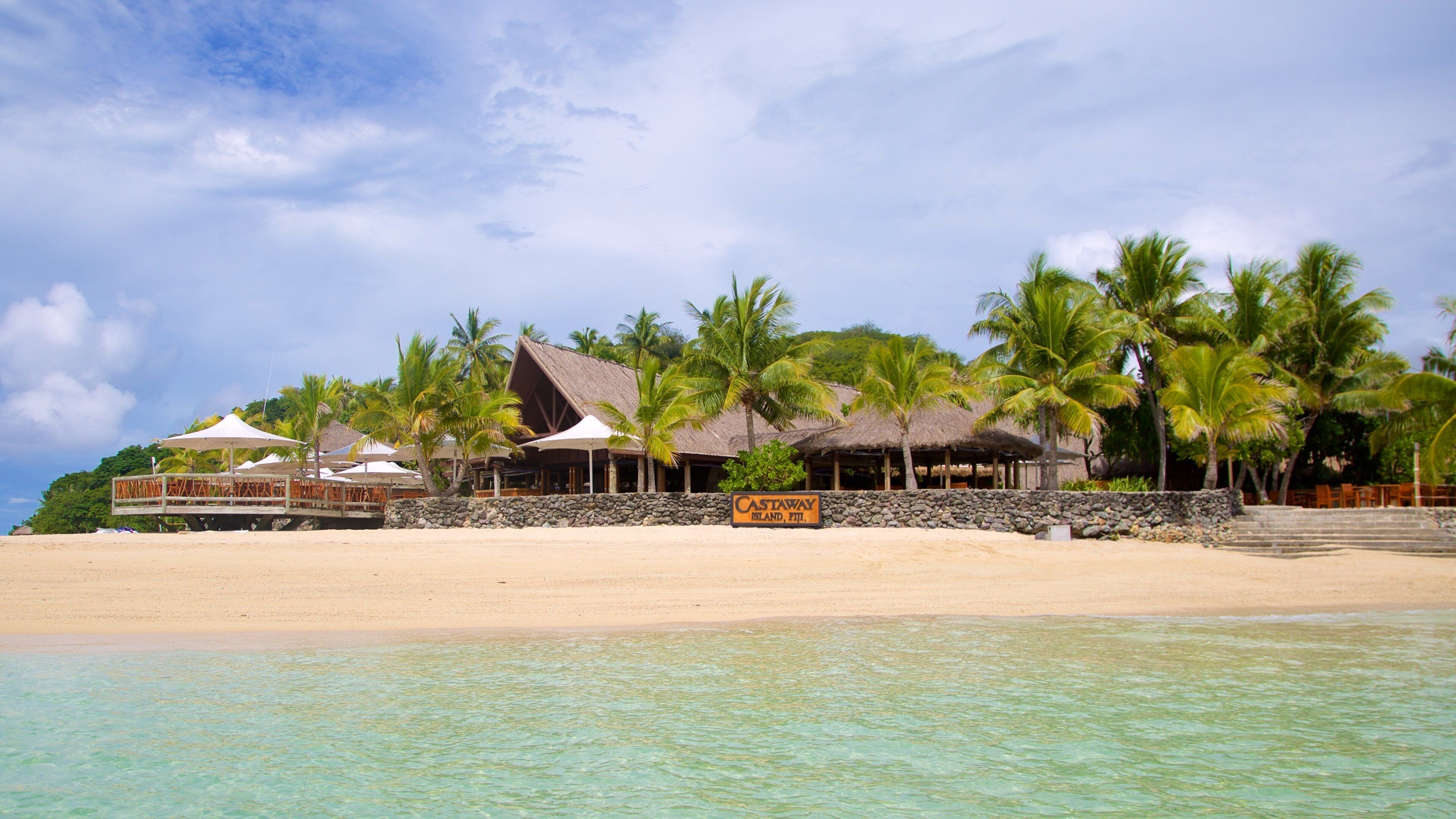 A beach resort with thatched-roof buildings, palm trees, and clear turquoise water.