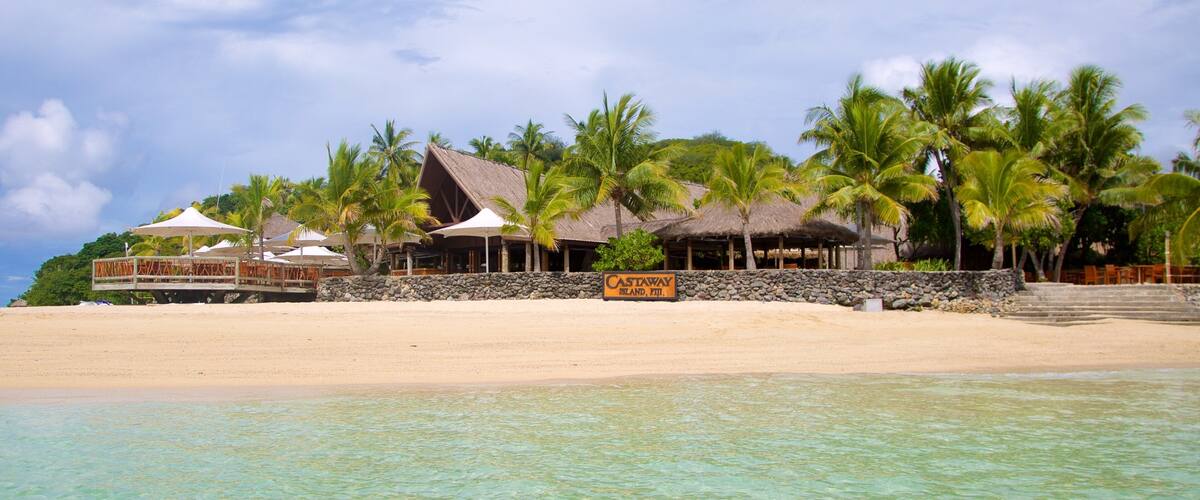 A beach resort with thatched-roof buildings, palm trees, and clear turquoise water.