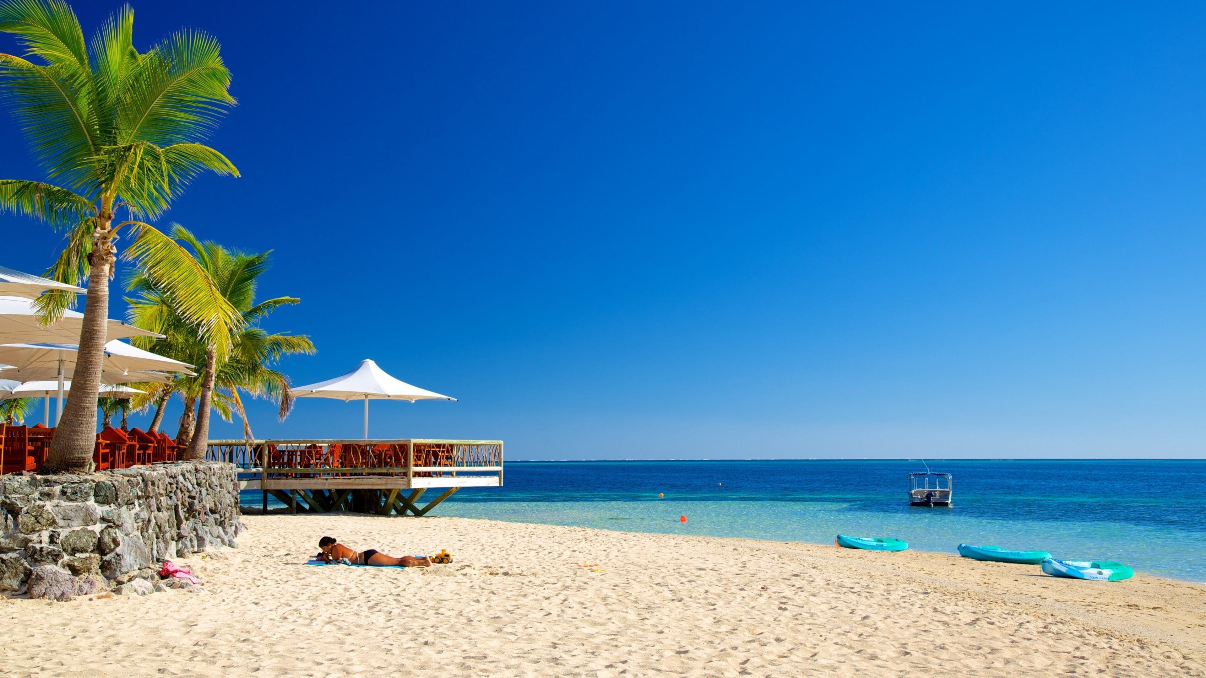 A beach with palm trees, a wooden structure with umbrellas, and a clear blue sky.