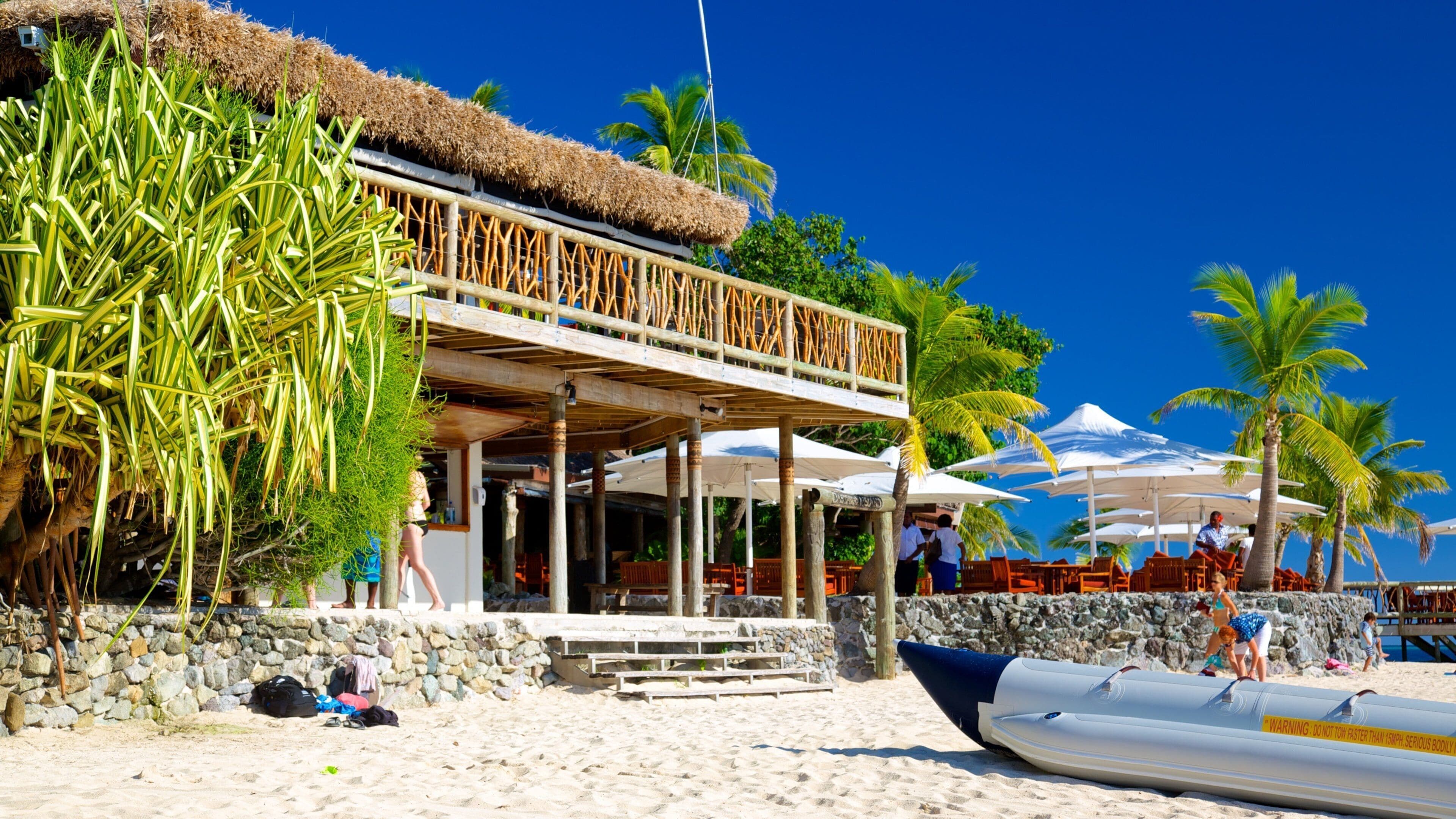 A beachside restaurant with a thatched roof, palm trees, and a lifeguard stand.