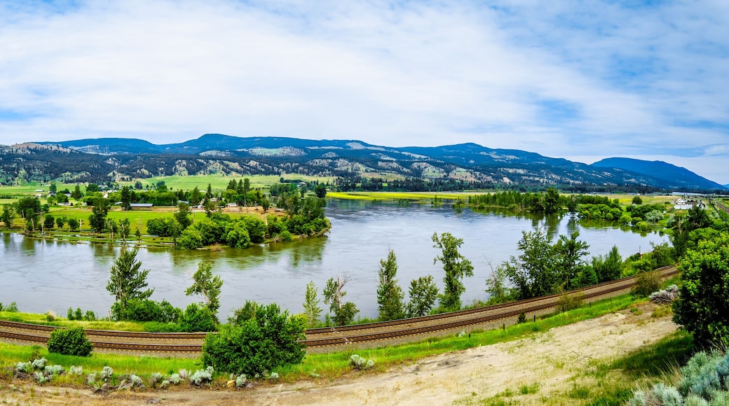 A river with a railway track curving around it, surrounded by green fields and mountains in the background.